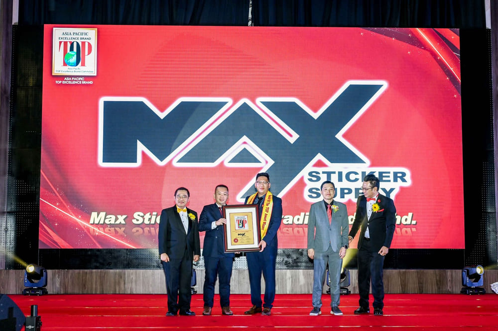 An award presentation on stage in front of a red LED wall carrying the MAX STICKER SUPPLY wordmark and the line "Max Sticker Supply Trading Sdn. Bhd."; one person holds a framed certificate.