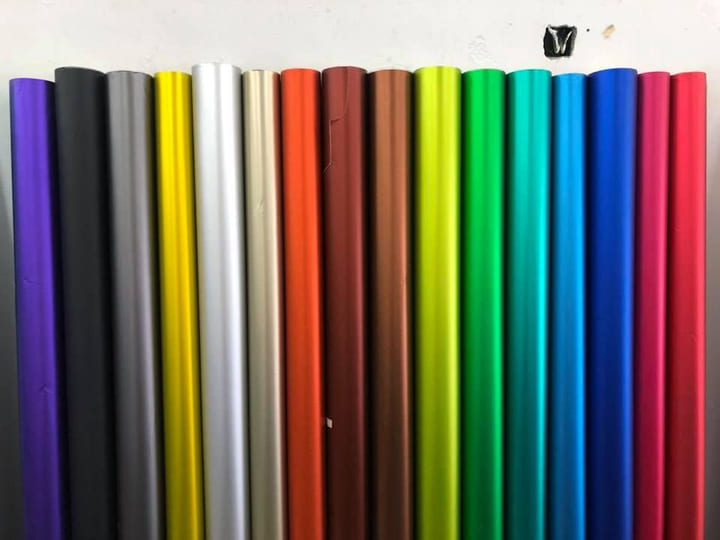 A line of vinyl roll ends stood against a white wall, showing around twenty film colours side by side from black and grey through reds, yellows, greens and blues.