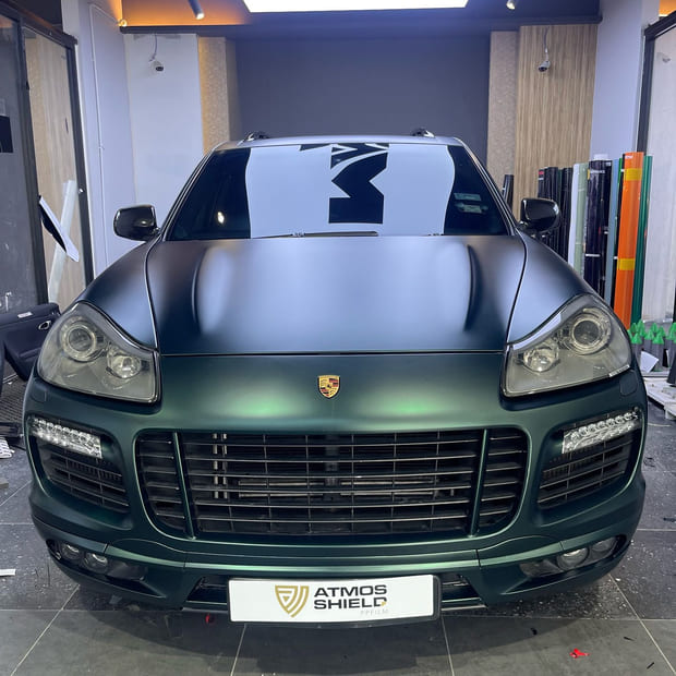 A Porsche Cayenne head-on in a bay with a pale wood-panelled wall, finished in matte metallic dark green.