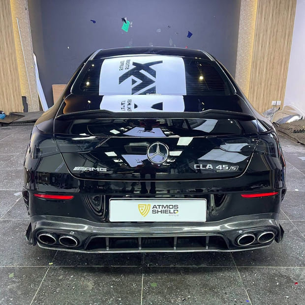 A Mercedes-AMG CLA 45 S from directly behind on the workshop floor, in gloss black film, with a carbon diffuser and quad tailpipes.