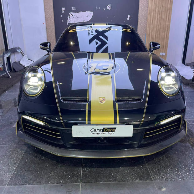 A Porsche 911 Turbo S head-on inside the Puchong workshop with its headlights lit, in gloss black film with a gold centre stripe and gold pinstriping, on gold wheels.