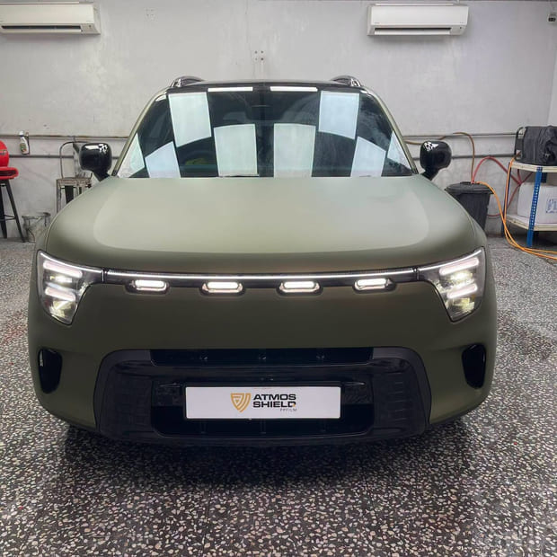 A smart #5 SUV head-on in the workshop in a matte olive green film, with a protective chequered sheet still over the windscreen.